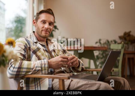 Image of handsome businessman working from cafe, drinking coffee and doing online project on his laptop, smiling and looking hap Stock Photo