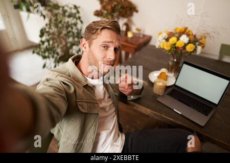 Handsome and confident young man taking selfie on mobile phone, pointing finger at camera, sitting in cafe with laptop, working Stock Photo