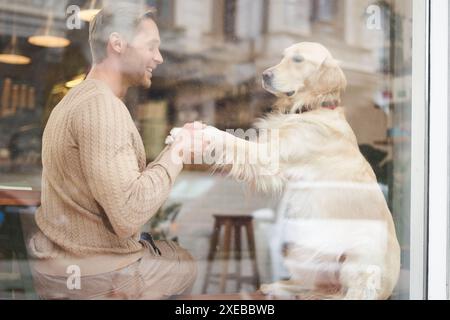 An outdoor shot of a man sitting with his dog in a pet-friendly cafe near window, golden retriever gives paw to visitor Stock Photo