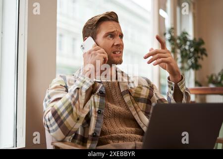 Image of man feeling stressed while talking over the phone. Businessman sitting in cafe with laptop, answering a call, hear bad Stock Photo
