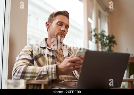 Portrait of man with serious face, scrolling news feed on mobile phone, looking concentrated at smartphone screen, sitting in ca Stock Photo