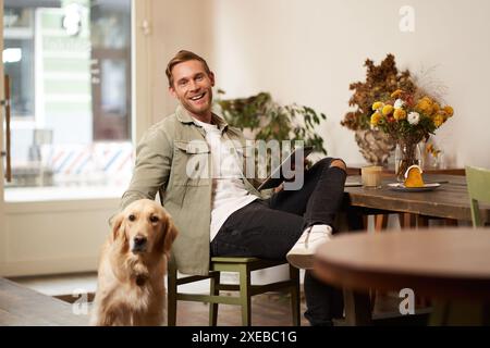 Portrait of handsome young man, cafe visitor, sitting with his dog, petting golden retriever and reading news on digital tablet, Stock Photo