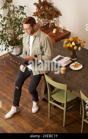 Portrait of handsome young man sits with laptop, looks at screen with concentrated, thoughtful face, working on a project, e-lea Stock Photo