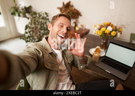 Handsome smiling young man taking selfie, showing okay good hand sign, photographing on mobile phone, sits in cafe with laptop Stock Photo