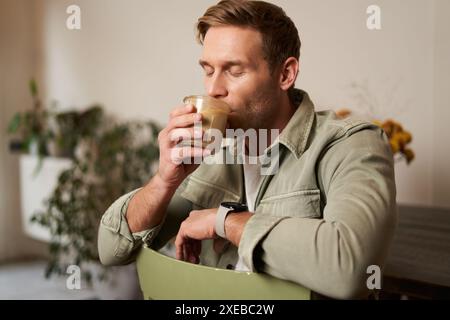 Cafe, leisure and lifestyle concept. Portrait of handsome young man sits on chair with glass of coffee, drinks his beverage and Stock Photo