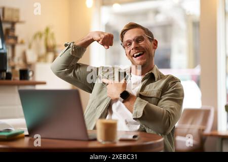 Portrait of handsome, pleased young man shows biceps, flexing muscles, sits in front of laptop in cafe, rejoicing, private coach Stock Photo