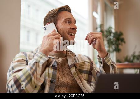 Portrait of young businessman, freelancer working remotely from coffee shop in city, sitting in chair with laptop, answer a phon Stock Photo