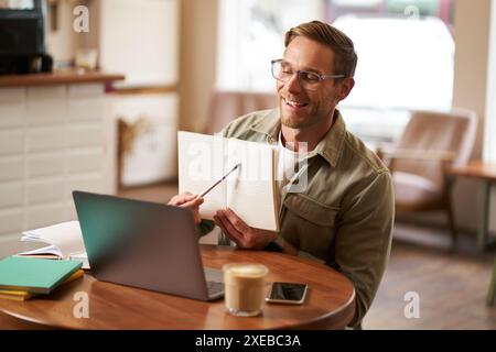 Portrait of handsome man in glasses, working remotely from empty cafe, showing example in his notebook, tutoring, using laptop t Stock Photo