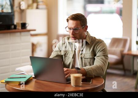 Portrait of man with serious face, sitting in cafe, wearing glasses, working on laptop, looking concentrated, doing freelance pr Stock Photo