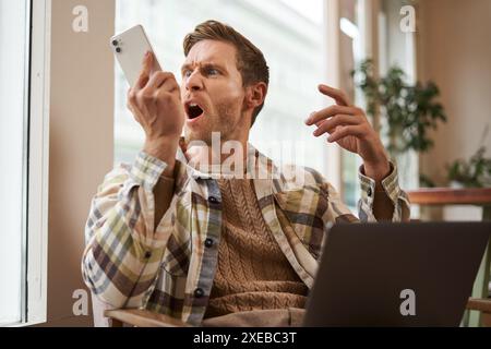 Portrait of an angry man shouting at his mobile phone. Frustrated cafe visitor arguing with someone on call, sitting with laptop Stock Photo