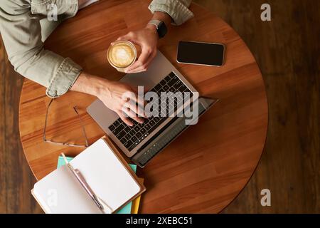 Co-working space, freelance and e-learning concept. Top view, male hands holding cup of coffee and using laptop. A man sitting i Stock Photo
