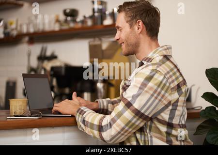 Portrait of handsome smiling man, sitting with laptop in a cafe, typing, working remotely from co-working space, drinking coffee Stock Photo