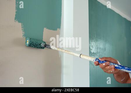 Anonymous male hand while standing in room painting green color on wall with help of roller brush attached to tip of metal rod in day time during reno Stock Photo