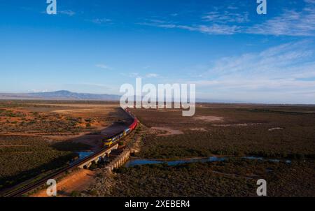 Aerial view of a freight train in motion, traveling through the outback near Port Augusta. The train is on an elevated track, crossing a bridge over a Stock Photo