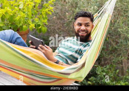 Relaxing in hammock, man using tablet and smiling outdoors in garden Stock Photo