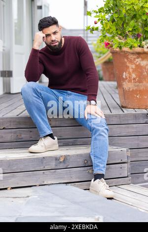 Outside his home, Man sitting on wooden steps, posing thoughtfully, wearing casual clothes Stock Photo