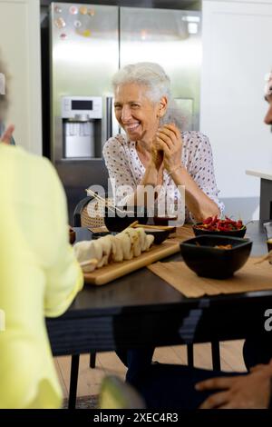 At home, Smiling senior woman enjoying meal with friends at dining table in kitchen Stock Photo