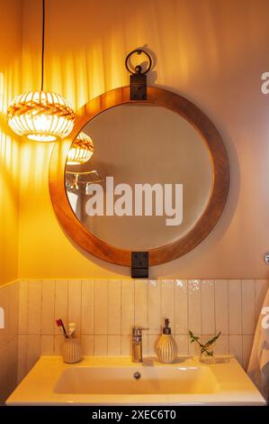 A round mirror in a wooden frame and a lamp with a wicker shade over the washbasin. A close-up of a part of the bathroom interio Stock Photo