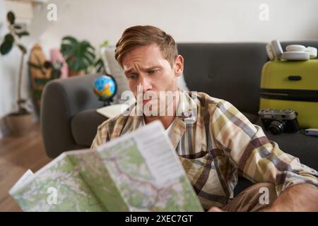 Portrait of man with concerned, puzzled face looking at map, frowning while studying the route, sitting with suitcase, going on Stock Photo