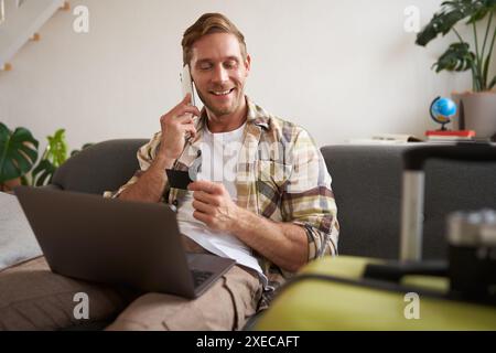 Image of guy calling someone, holding credit card and using laptop, has suitcase and camera packed for holiday Stock Photo