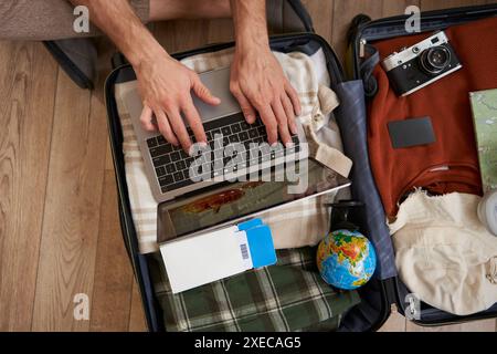 Close up of tourist, hands typing on laptop, opened suitcase with clothes for holiday, man packs luggage Stock Photo