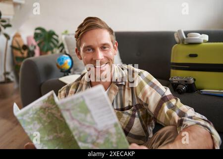 Portrait of handsome smiling man, holding map, sitting with suitcase in living room, going abroad on holiday, studying his route Stock Photo