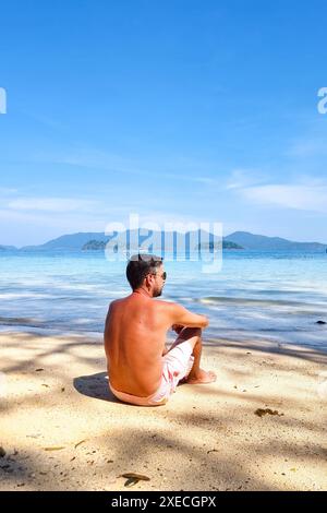 A man in casual clothing sits on the sandy beach, his gaze fixed out to the vast expanse of the ocean before him Stock Photo