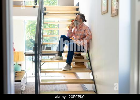 Sitting on stairs, senior man drinking coffee and looking out window at home Stock Photo