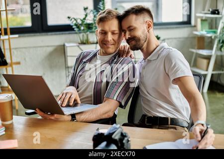 Two men in casual attire sitting at table, engrossed in work on laptop. Stock Photo