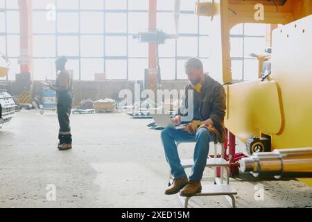 Wide shot of Caucasian technician using laptop to change settings of industrial machine, his Black colleague using digital tablet in background Stock Photo
