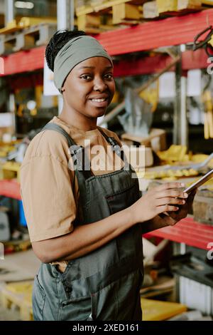 Vertical medium portrait of cheerful Black female specialist working in factory warehouse smiling at camera while using digital tablet Stock Photo