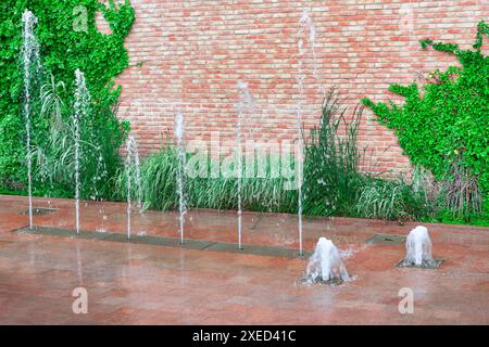 Wall with a brick facade and a row of fountains. The fountains are spraying water over the pavement Stock Photo
