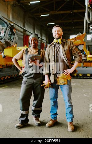 Vertical long shot of cheerful ethnically diverse male and female engineers posing for camera at work in machinery production factory Stock Photo