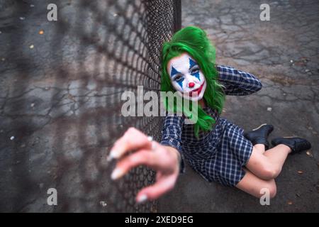 Greenhaired girl in chekered dress with joker makeup sitting near wire mesh fence on the ground. Wide shot. Stock Photo