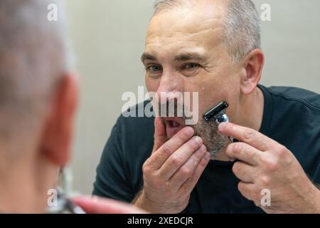 Mature gray-haired man restyling his beard himself at home using razor Stock Photo