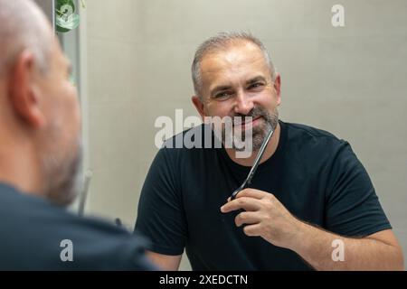 Portrait of smiling Middle-aged handsome man cutting his beard with scissors. Selfcare during quarantine isolation Stock Photo