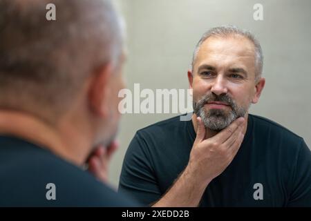 Middle-aged handsome smiling man looking in mirror in bathroom touching his beard thinking of cutting it off Stock Photo