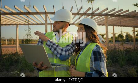 Two Caucasian experienced engineers workers woman female girl man male guy hardhat using laptop pc gadget device computer discuss talking project Stock Photo