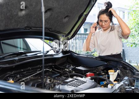 A woman using her phone to talk about her broken car. She has a worried expression on her face and is explaining the car's problems to the person on t Stock Photo