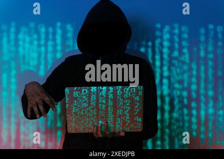 A hacker is sitting in a dark room, hunched over a laptop computer. The hacker is wearing a hoodie and sunglasses, and their face is hidden in the sha Stock Photo