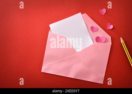 Blank white paper in a pink envelope on a red background with pink hearts and pencil. Top view. Stock Photo