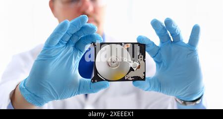 A male repairman wearing blue gloves is holding a hard drive from computer or laptop in hands. Performs fault diagnostics and performs urgent repairs Stock Photo