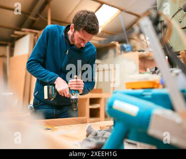 Carpenter At Workbench In Workshop Using Cordless Electric Drill Stock Photo