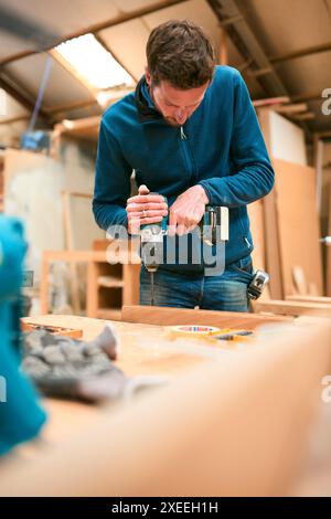 Carpenter At Workbench In Workshop Using Cordless Electric Drill Stock Photo