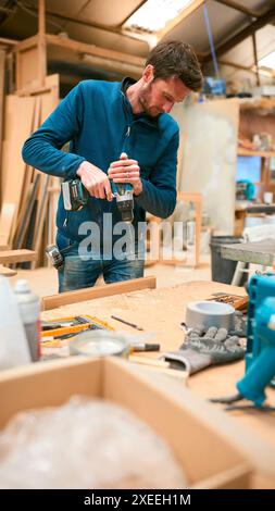 Carpenter At Workbench In Workshop Using Cordless Electric Drill Stock Photo