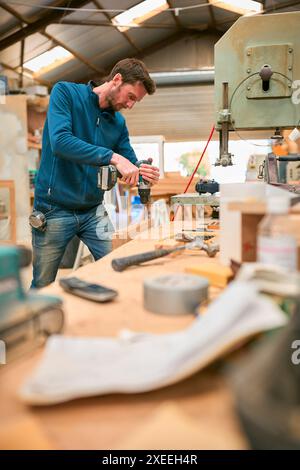 Carpenter At Workbench In Workshop Using Cordless Electric Drill Stock Photo