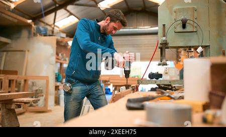 Carpenter At Workbench In Workshop Using Cordless Electric Drill Stock Photo
