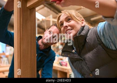 Carpenter With Female Apprentice In Workshop Using Ruler To Measure Wood Of Window Frame Stock Photo