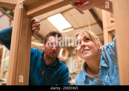 Carpenter With Female Apprentice In Workshop Using Ruler To Measure Wood Of Window Frame Stock Photo