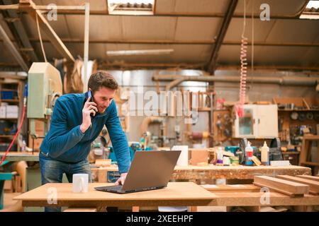 Carpenter Working In Woodwork Workshop Talking On Mobile Phone And Using Laptop Stock Photo